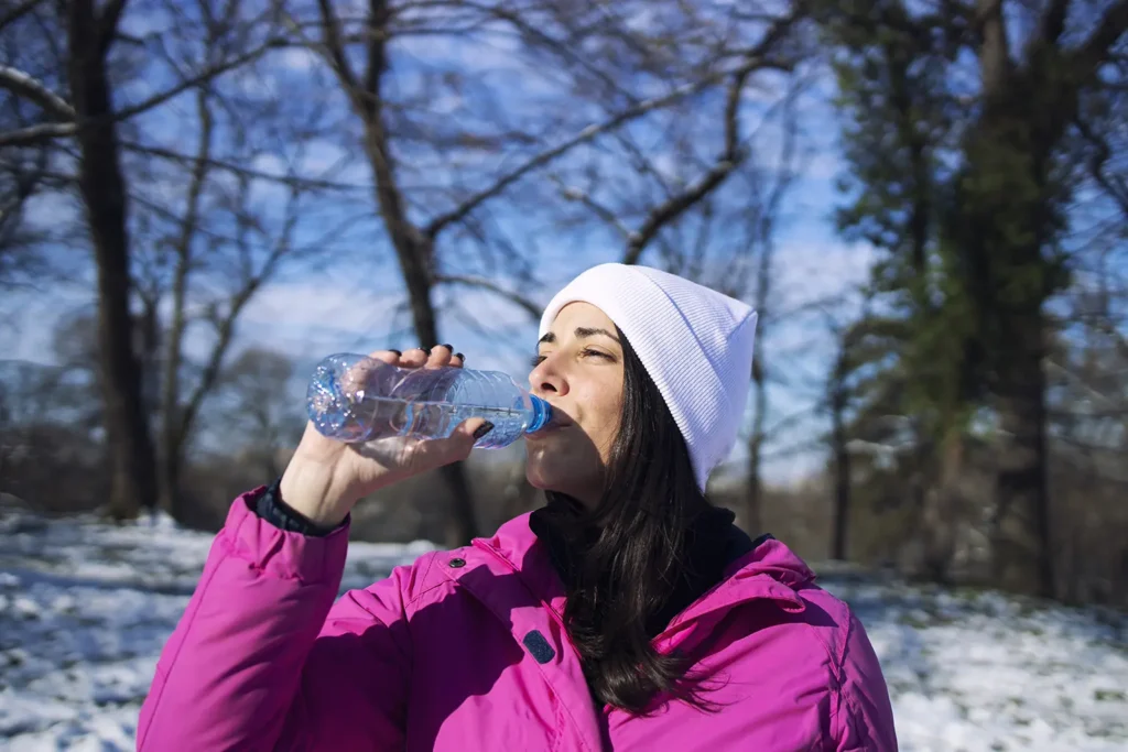 Female athlete in winter clothes drinking water outdoors, showing how cold weather can affect your teeth and the necessity of staying hydrated to maintain oral health during winter.