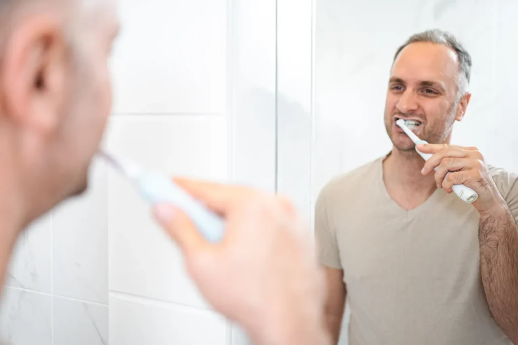 Man brushing his teeth with an electric toothbrush in front of a bathroom mirror, highlighting the benefits of using an electric toothbrush for maintaining good oral hygiene.