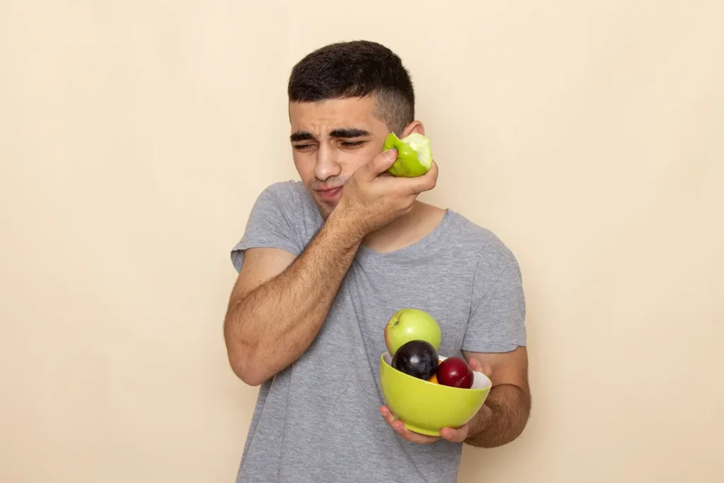 Man experiencing teeth sensitivity while biting an apple, holding his jaw in pain, illustrating why my teeth hurt.